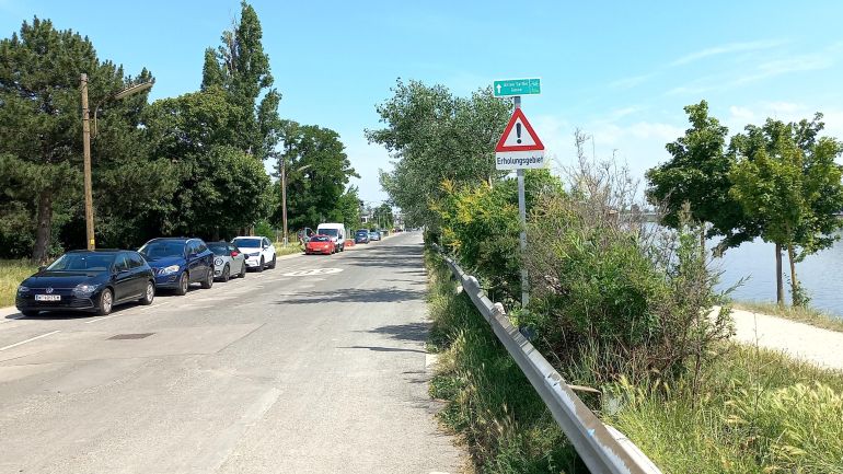 copyright: MA 46 Rechts neben der Fahrbahn ist ein Hinweisschild für einen Radweg, darunter "Achtung" mit Zusatz "Erholungsgebiet". Auf einer Straßenseite parken Autos.