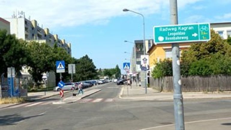 copyright: MA 46 An einer Kreuzung hängt ein Schild, das einen Radweg nach rechts in Richtung Kagran anzeigt. Geradeaus ist ein Zebrastreifen. Es fahren keine Autos.