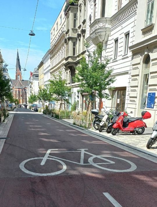 Auf einer Straße ist ein großes Fahrradsymbol aufgezeichnet. Am Straßenrand parken mehrere Mopeds zwischen Bäumen.