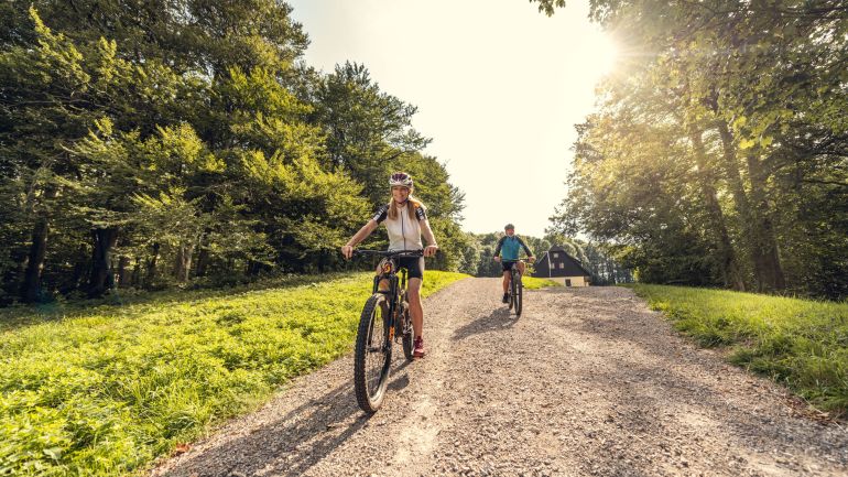 Frau und Mann fahren mit Mountainbikes auf einem befestigten Weg, links und rechts Wald