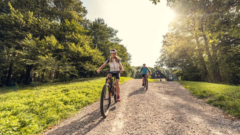 Frau und Mann fahren mit Mountainbikes auf einem befestigten Weg, links und rechts Wald