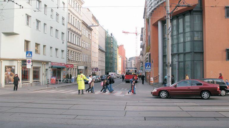 Kreuzungsbereich Quellenstraße - Wielandgasse: Kreuzung mit rot unterlegtem Zebrastreifen und einem Schülerlotsem