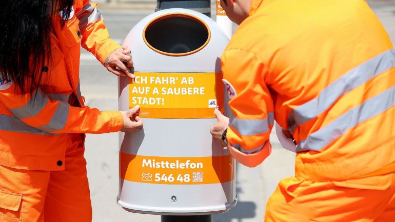 copyright: Stadt Wien/Martin Votava 2 Mitarbeiter*innen der MA 48 bekleben einen Mistkübel mit dem Sticker "Ich fahr' ab auf a saubere Stadt"