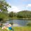 Am Ufer eines ruhigen Gewässers sitzt eine Familie in Badekleidung auf einer Picknickdecke. Die Kinder haben einen aufblasbaren Delfin dabei.