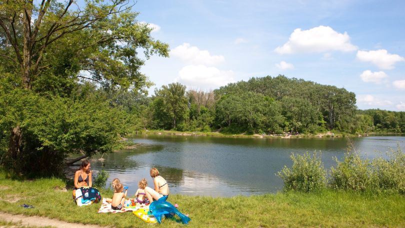 Am Ufer eines ruhigen Gewässers sitzt eine Familie in Badekleidung auf einer Picknickdecke. Die Kinder haben einen aufblasbaren Delfin dabei.
