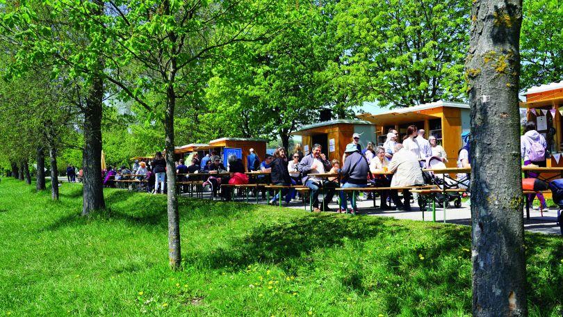 Leute sitzen im Grünen vor Verkaufsständen auf Bierbänken