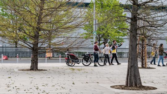 Mehrere Menschen mit Fahrrädern vor einem Glaskomplex, davor Bäume