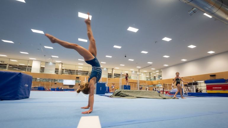 Junge Frau macht Handstand, im Hintergrund Sportgeräte und weitere junge Frauen.