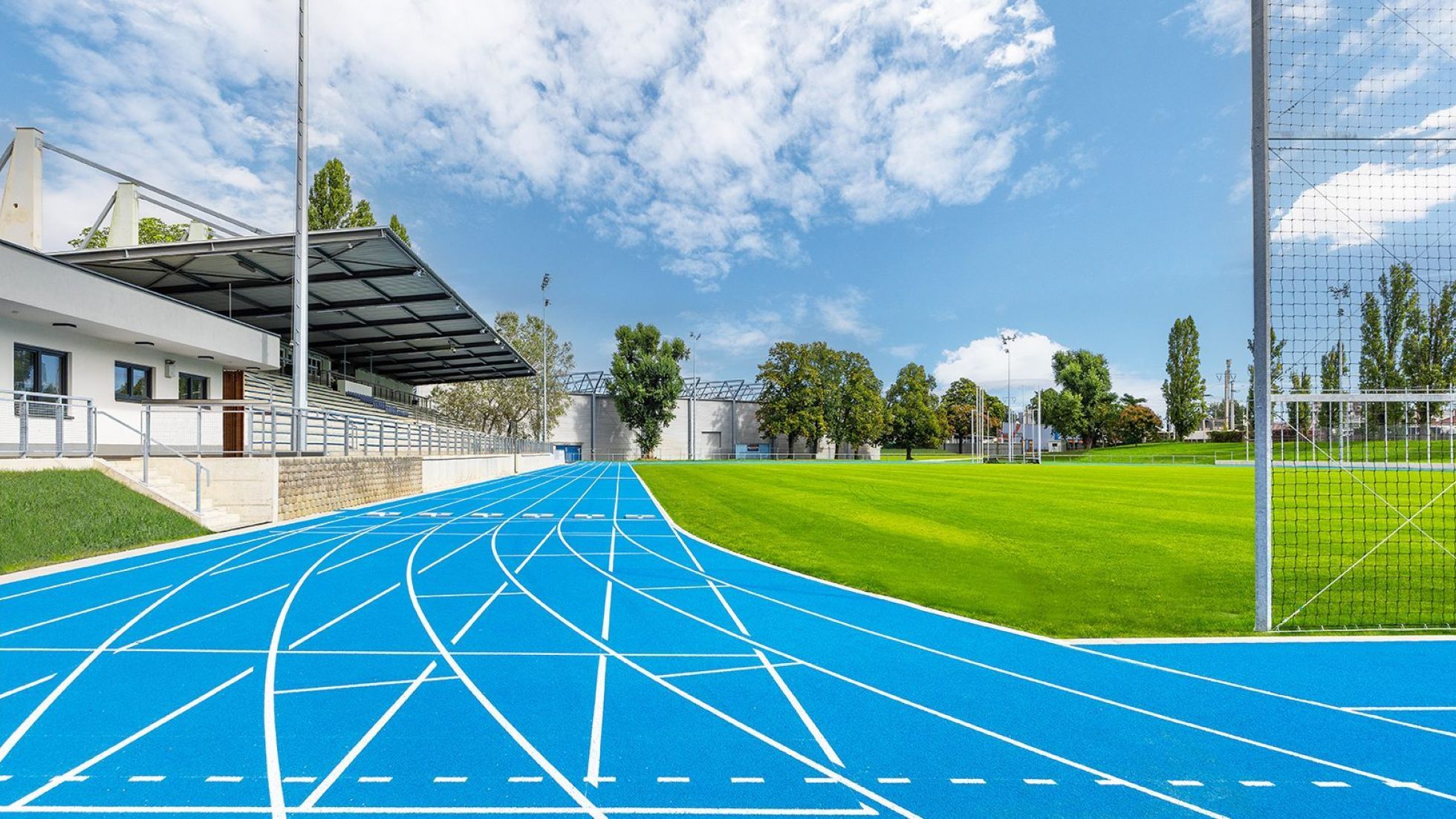 Outdoor-Sportanlage mit blauer Laufbahn, grünem Rasen in der Mitte und einer Tribüne am Rand der Bahn.