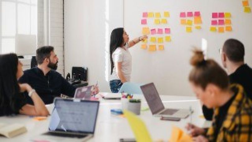 copyright: unsplash - you x ventures Eine Frau erklärt etwas an einem Whiteboard, eine Gruppe von Personen an einem Tisch folgt den Erläuterungen.
