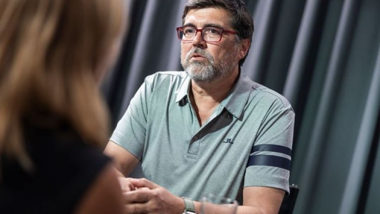 Frau interviewt Mann mit grauem Polo-Shirt, roter Brille, grauem Bart und Kurzhaarfrisur