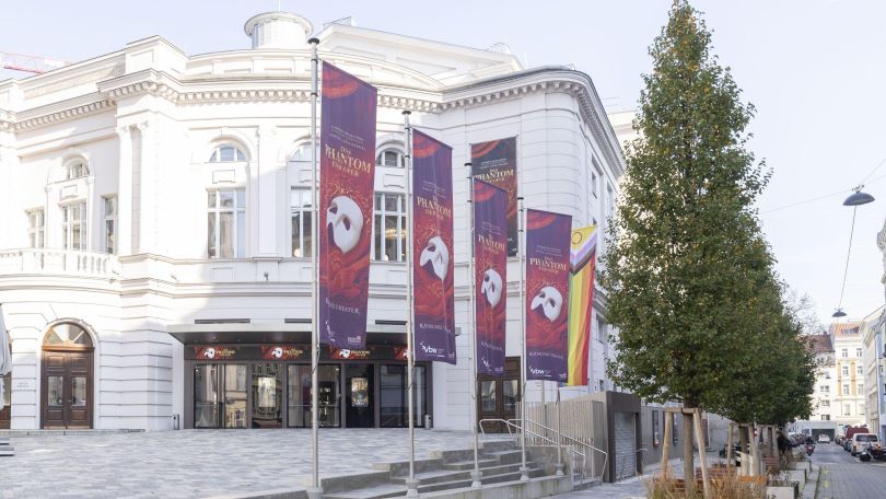 Vor einem historischen Theatergebäude mit heller Fassade wehen Fahnen für das Musical "Das Phantom der Oper".