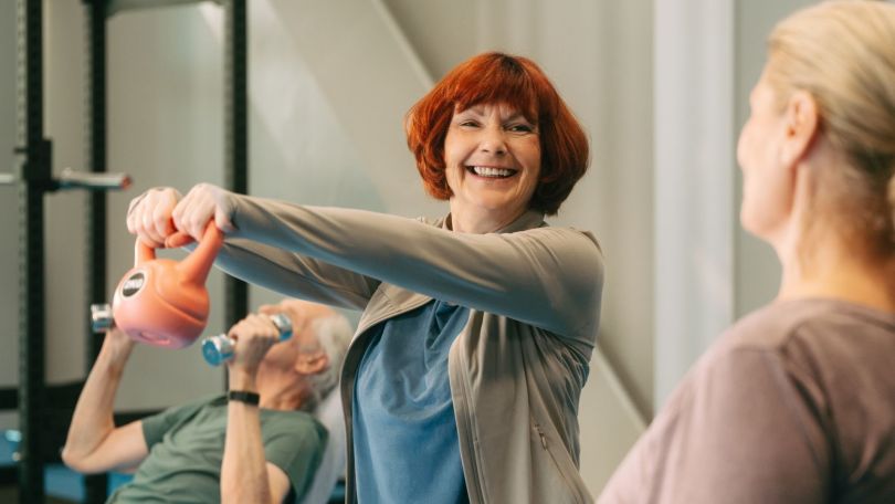 copyright: Jacky Zoe - Stadt Wien Seniorin mit roten kinnlangen Haaren schwingt Kettlebell, lacht einer anderen Seniorin zu, im Hintergrund trainiert ein Senior mit kleinen Hanteln.