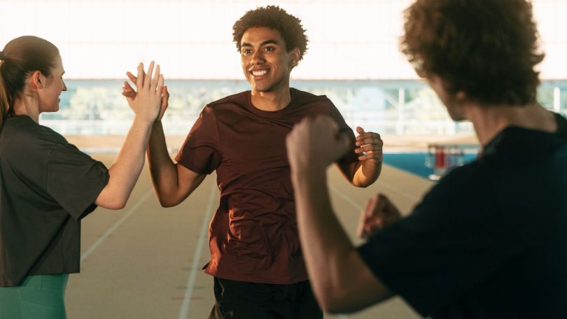 Drei Personen auf einer Laufstrecke: Ein Mann lächelt und gibt einer Frau ein High-Five, während eine andere Person zusieht.