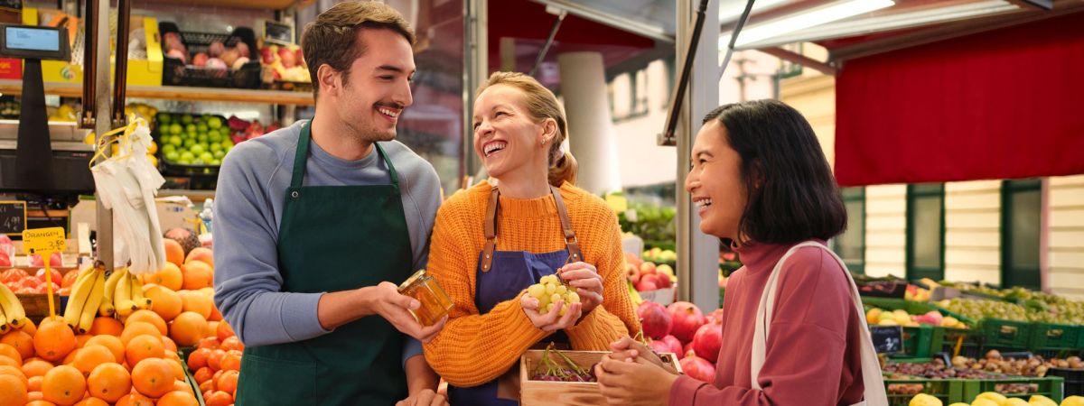 copyright: Stadt Wien/Kerstin Hammerschmid 3 lachende Personen stehen mit Produkten in der Hand vor einem Marktstand.