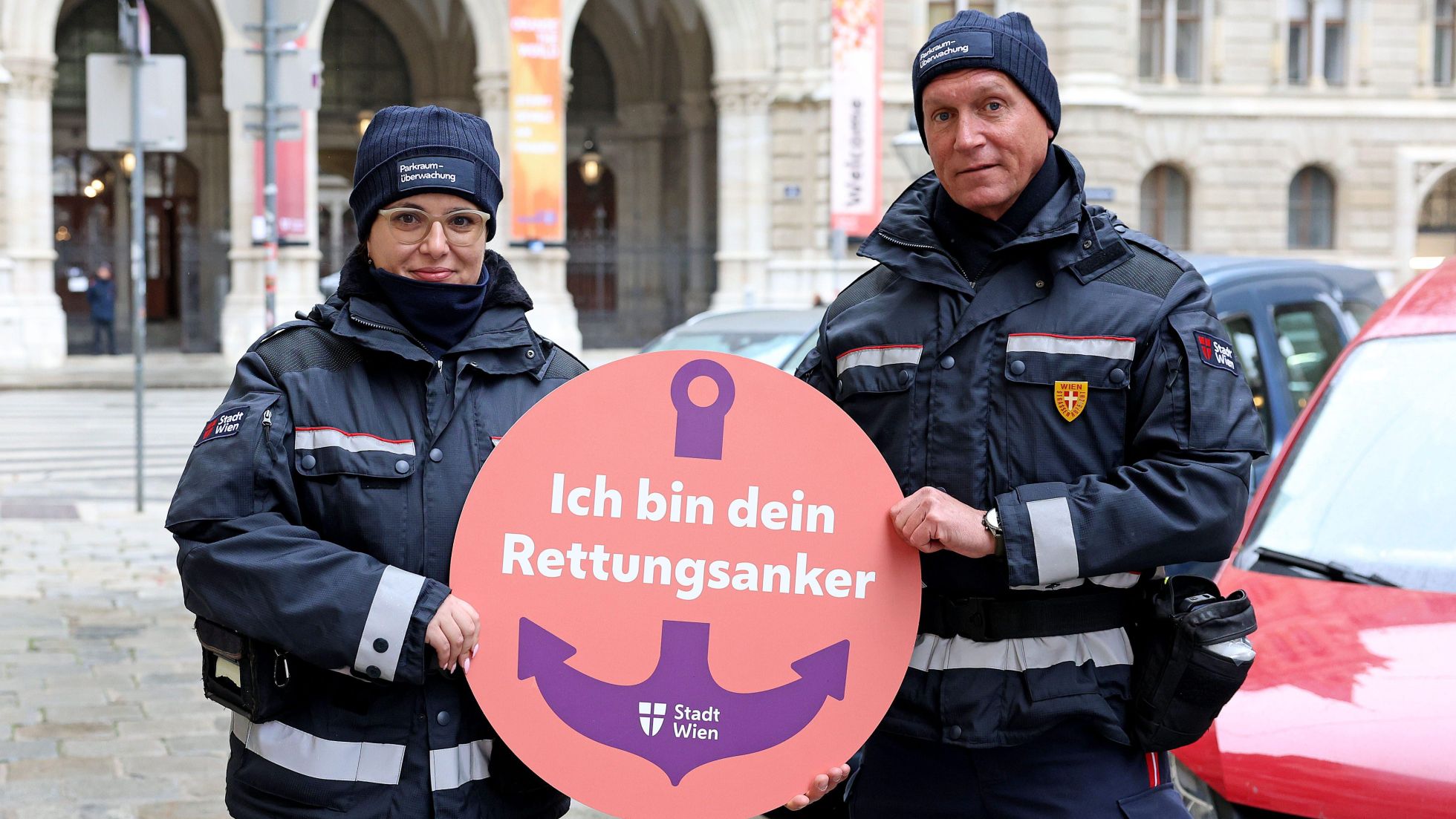 2 Mitarbeiter*innen der Parkraumüberwachung halten das Logo der Initiative "Ich bin dein Rettungsanker" in der Hand