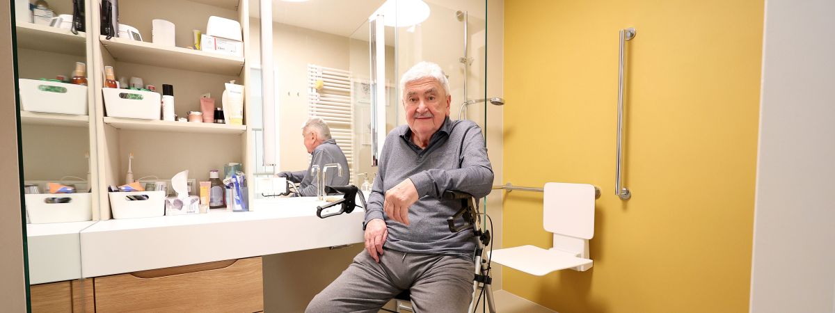 Ein Mann sitzt im Badezimmer auf seinem Rollator. In der Dusche sind ein Duschsitz und eine Haltestange montiert.