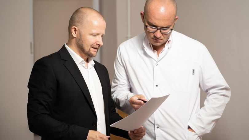 Arzt spricht mit einem Mann in schwarzem Sakko und zeigt auf Zettel