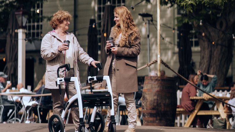 copyright: Pixabay Eine ältere Frau mit Rollator und eine jüngere Frau unterhalten sich und halten einen Kaffeebecher