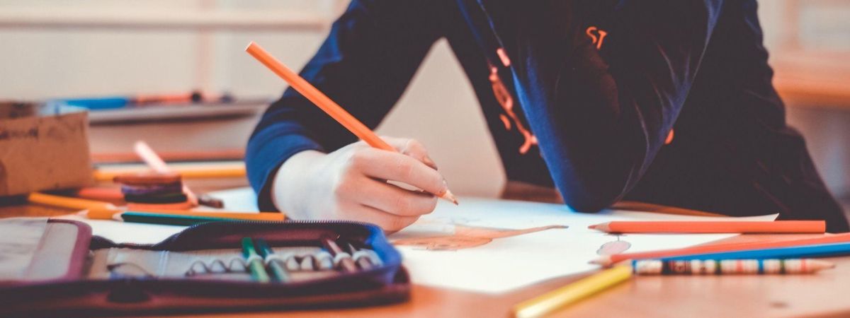 Bildausschnitt eines Schüler; man sieht seine Hand, die einen Stift über ein Schreibheft hält; am Schreibtisch liegen ein Federpennal und Stifte