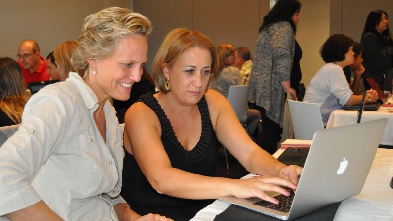 Ein Raum mit Seminarteilnehmer*innen; 2 Frauen sitzen vor einem Laptop