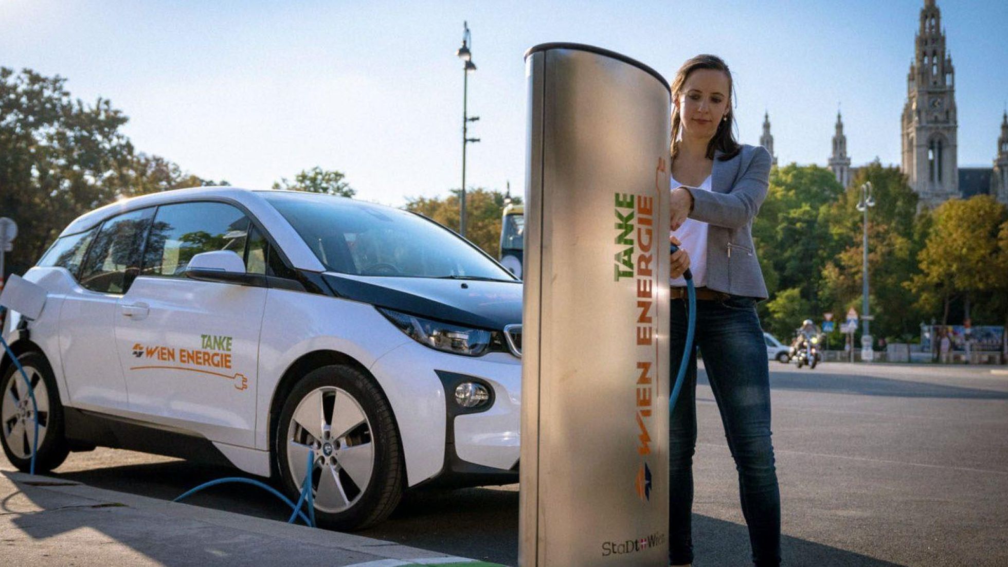 Eine Frau ladet ein weißes E-Auto von Wien Energie bei einer Ladestelle für E-Fahrzeuge auf. Im Hintergrund ist das Rathaus.