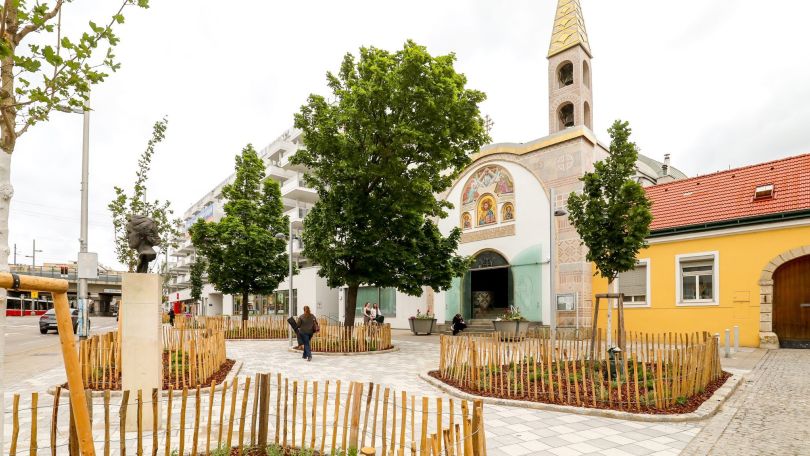 Platz vor einer Kirche in Simmering mit hellem Pflaster und großen, begrünten Baumscheiben.