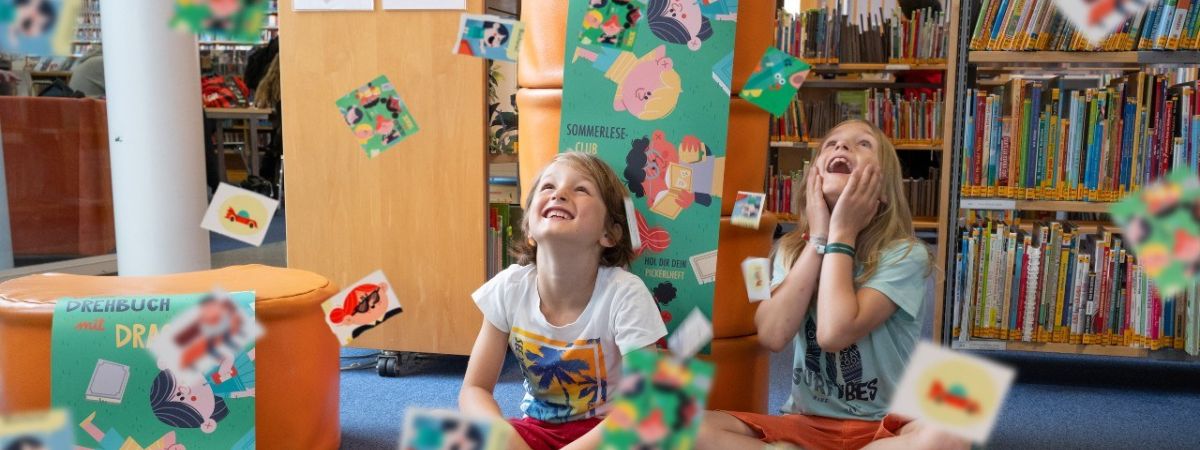 2 Kinder sitzen lachend auf dem Boden einer Bibliothek, während rund um sie zahlreiche bunte Sammelkarten durch die Luft fliegen. Im Hintergrund sind Bücherregale zu sehen.