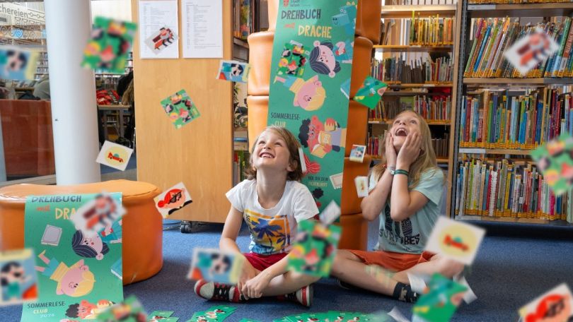 copyright: Stadt Wien Büchereien/Natalie Stephan 2 Kinder sitzen lachend auf dem Boden einer Bibliothek, während rund um sie zahlreiche bunte Sammelkarten durch die Luft fliegen. Im Hintergrund sind Bücherregale zu sehen.
