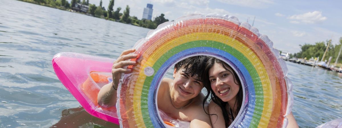 Ein junger Mann und eine junge Frau auf einer Luftmatratze schauen durch einen Schwimmring in Regenbogenfarben.