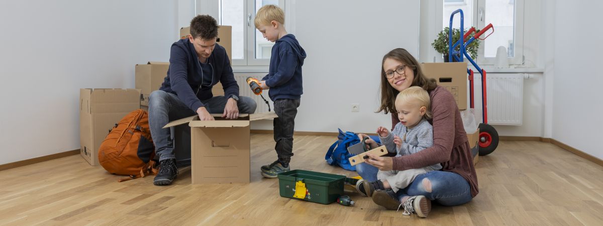 Mutter und Vater spielen mit ihren kleinen Söhnen in einem leeren Wohnraum und packen Umzugskisten aus.