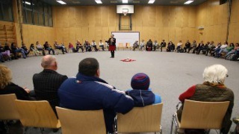 Personen in einem großen Sesselkreis hören einer Vortragenden zu