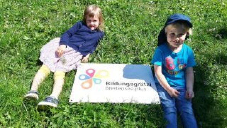 2 Kinder neben einer Bildungsgrätzl-Tafel in der Wiese