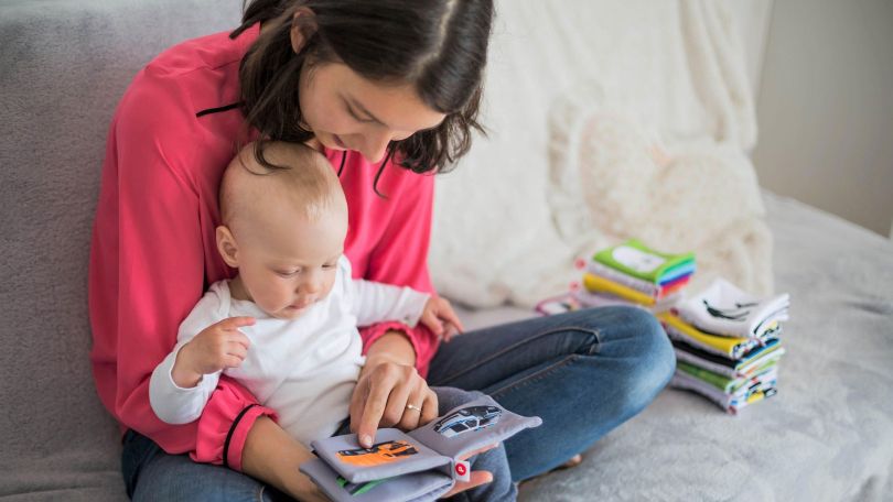 Eine junge Frau hält ein Baby auf dem Schoß und zeigt auf ein Bilderbuch