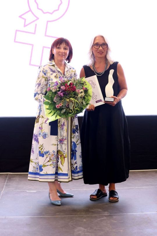 Eine Frau mit Blumenstrauss und eine Frau mit Urkunde und Auszeichnung in den Händen