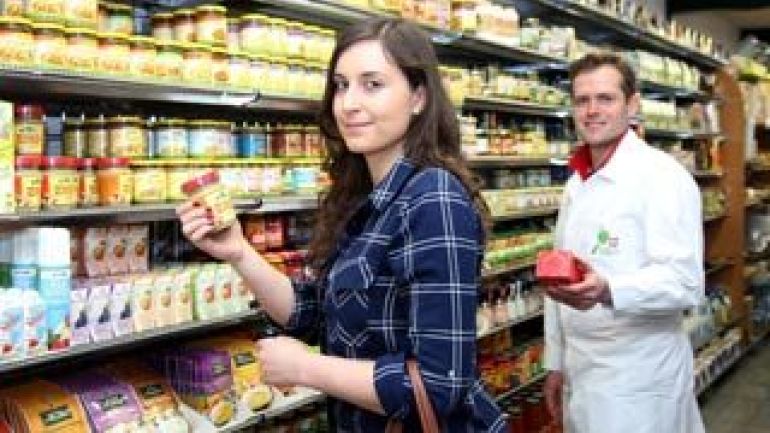 Frau in weißer Arbeitskleidung steht vor einem Supermarktregal und überprüft eine Packung