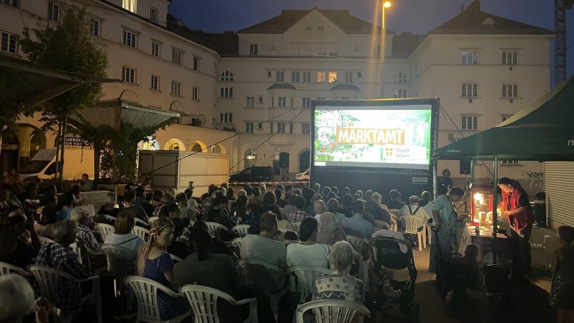 Viele Menschen sitzen auf weißen Plastiksesseln vor einer Freiluftkino-Leinwand in der Stadt.