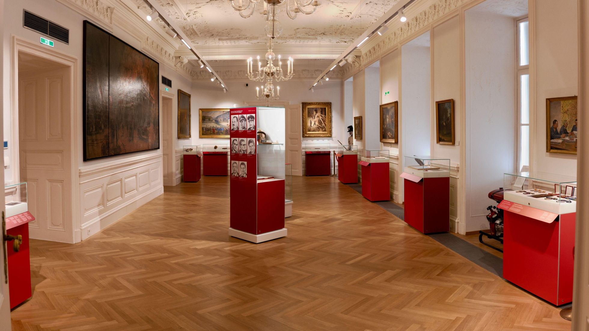 copyright: Stadt Wien - Feuerwehr Historischer Ausstellungsraum im Wiener Feuerwehrmuseum mit Parkettboden, Kronleuchtern und Gemälden an den Wänden. Mehrere rote Vitrinen präsentieren Exponate und Informationstafeln in einem hellen Museumsraum.