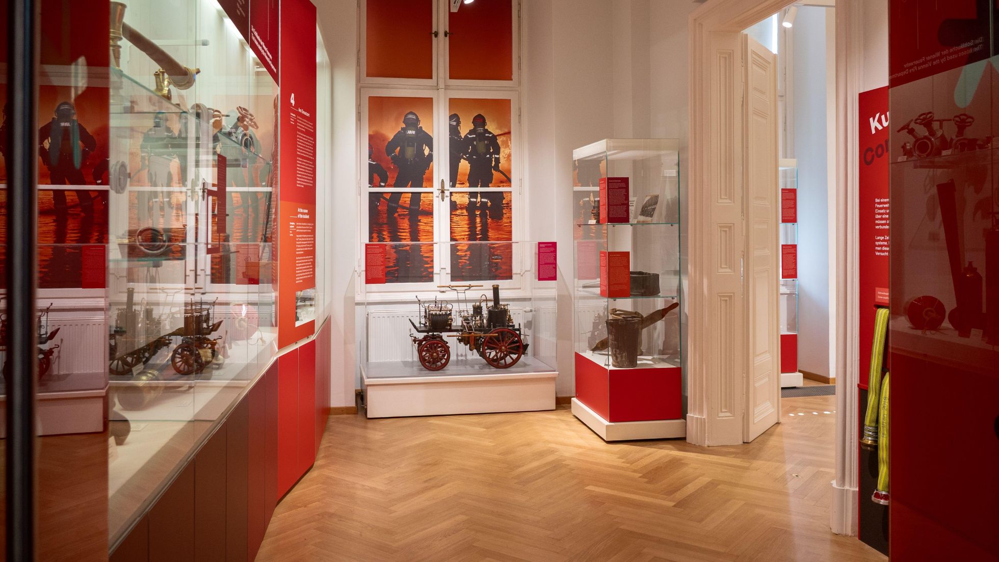 copyright: Stadt Wien - Feuerwehr Museumsraum im Wiener Feuerwehrmuseum mit roten Vitrinen und historischen Feuerwehr-Exponaten, darunter ein Modell eines Feuerwehrwagens. Im Hintergrund zeigen großformatige Bilder Einsatzkräfte bei einem Brandeinsatz.