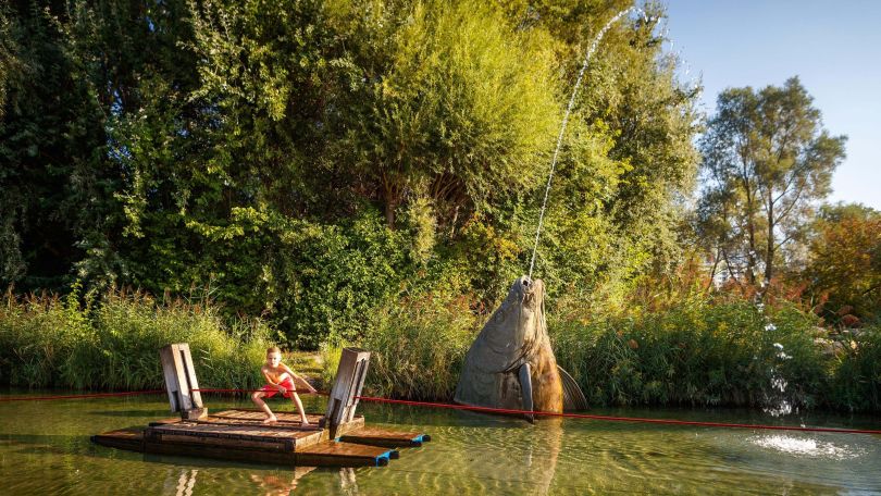 copyright: Wiener Stadtgärten - Houdek Ein Bub zieht sich an einem Seil mit einem Floß über seichtes Wasser. Im Hintergrund ist ein Springbrunnen in Form eines Fischkopfes.
