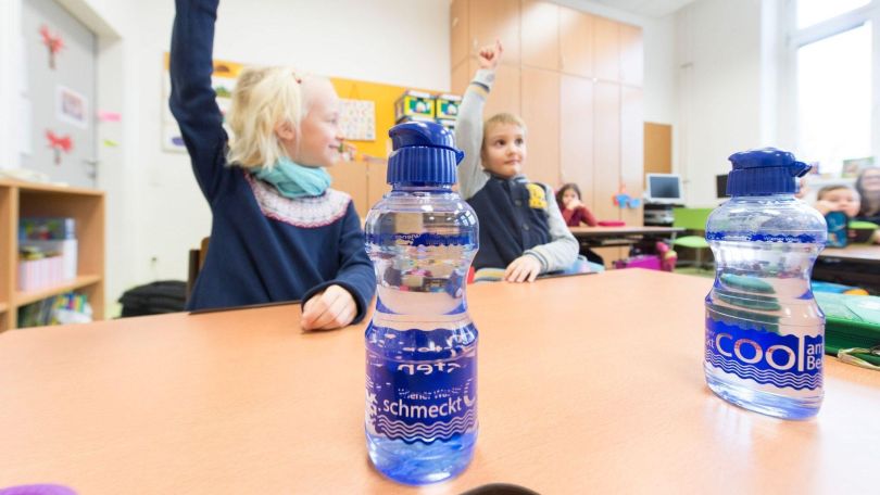 Kinder sitzen in einer Schulklasse und zeigen auf. Auf den Tischen stehen Wasserflaschen.
