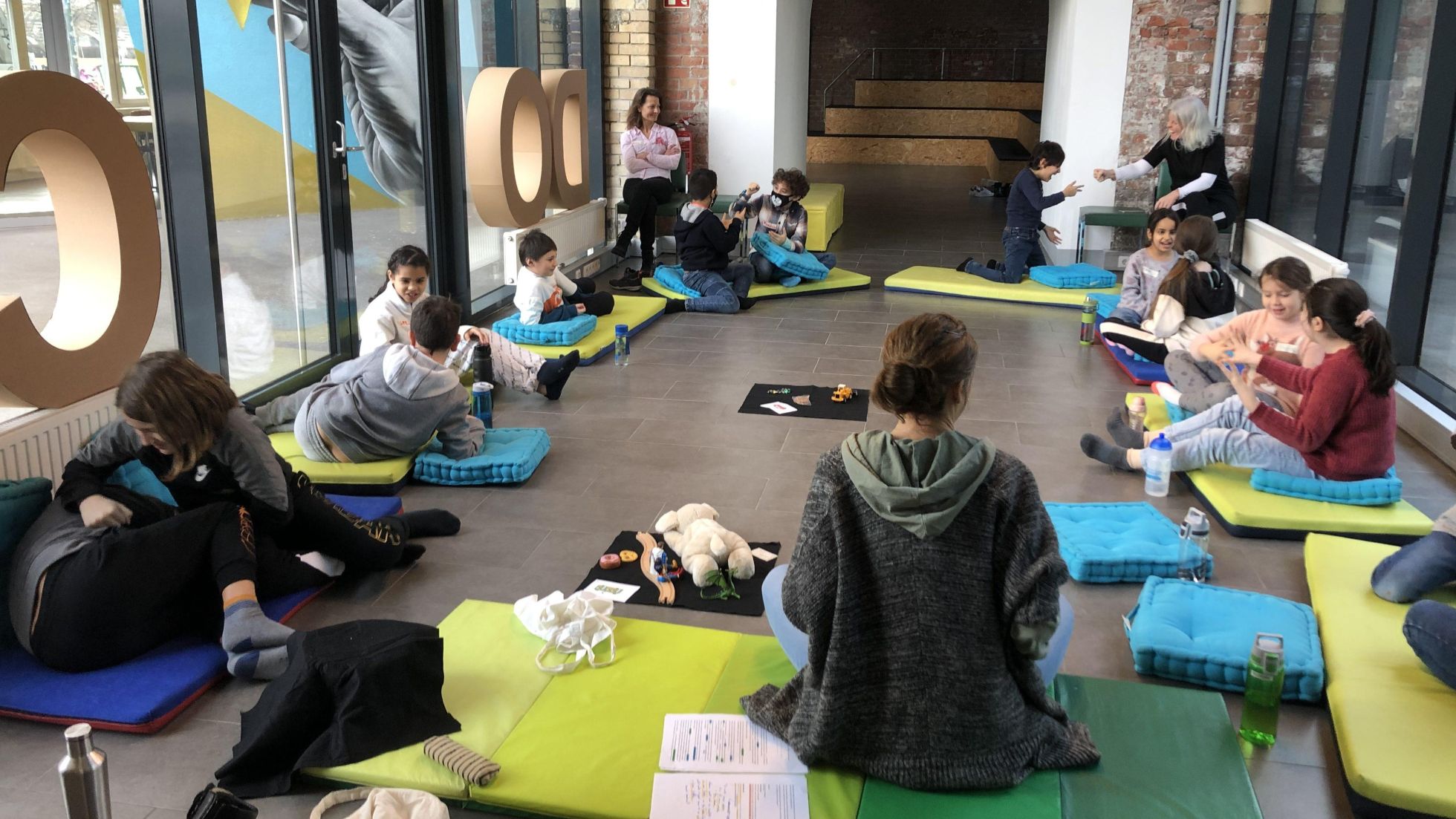 Eine Kindergruppe sitzt mit 2 erwachsenen Frauen auf gelben und blauen Matten am Boden