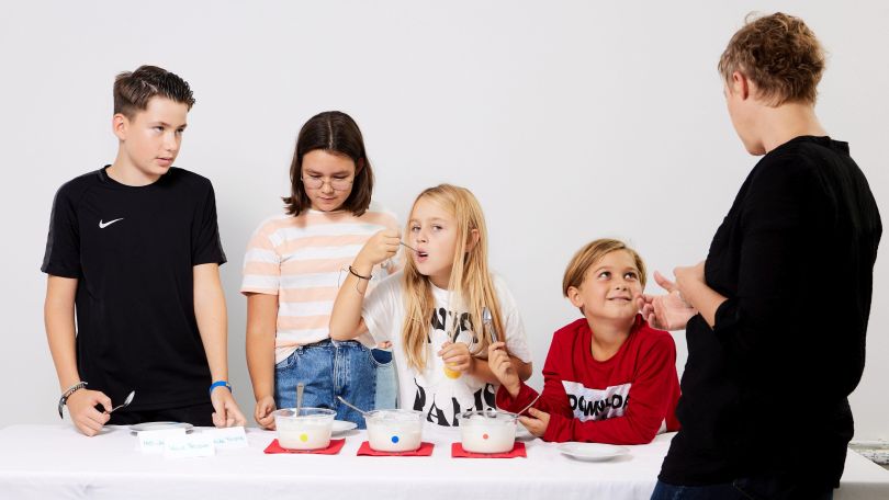 4 Kinder kosten Joghurt aus weißen Schüssel, vor dem Tisch der Kinder steht eine Frau mit dem Rücken zur Kamera