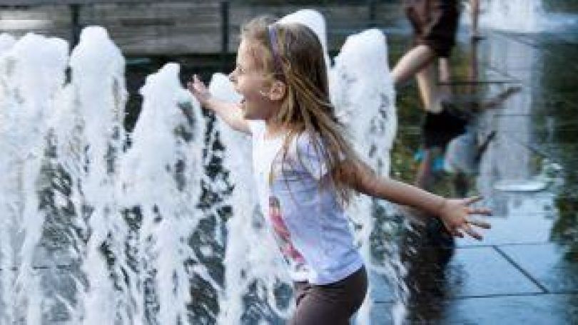 Mädchen läuft mit ausgebreiteten Armen vor einen Brunnen