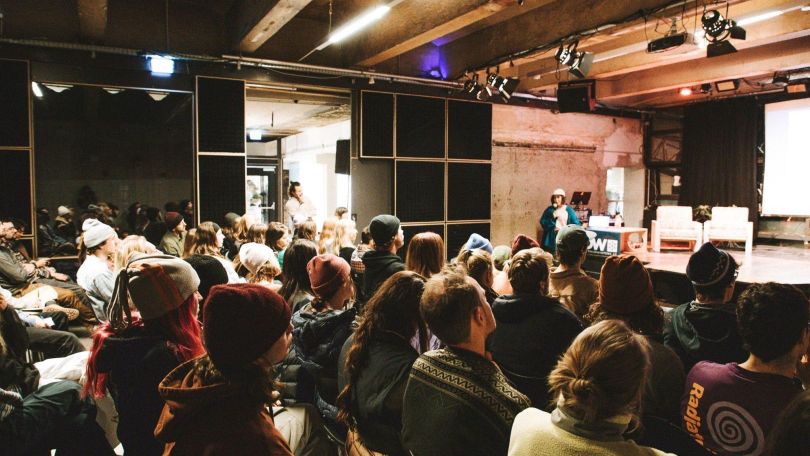 Junges Publikum sitzt bei einer Veranstaltung vor einer Bühne und hört einer Vortragenden zu.