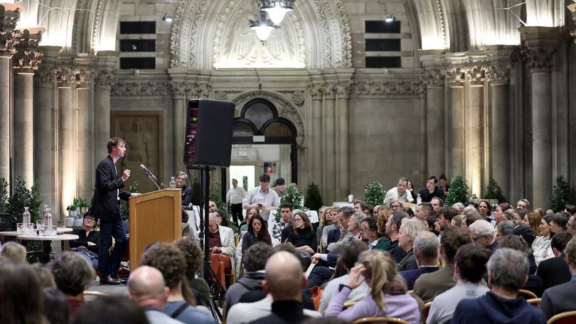 copyright: PID/VOTAVA Ein Mann am Rednerpult hält einen Vortrag vor einem großen Publikum in einem historischen Veranstaltungssaal.