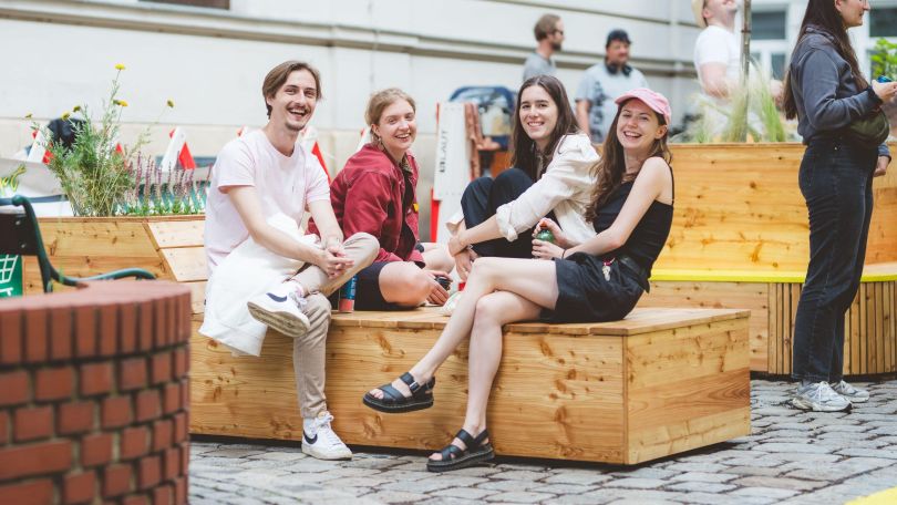 4 junge Menschen sitzen draußen auf einer Holzbank und lächeln in die Kamera.
