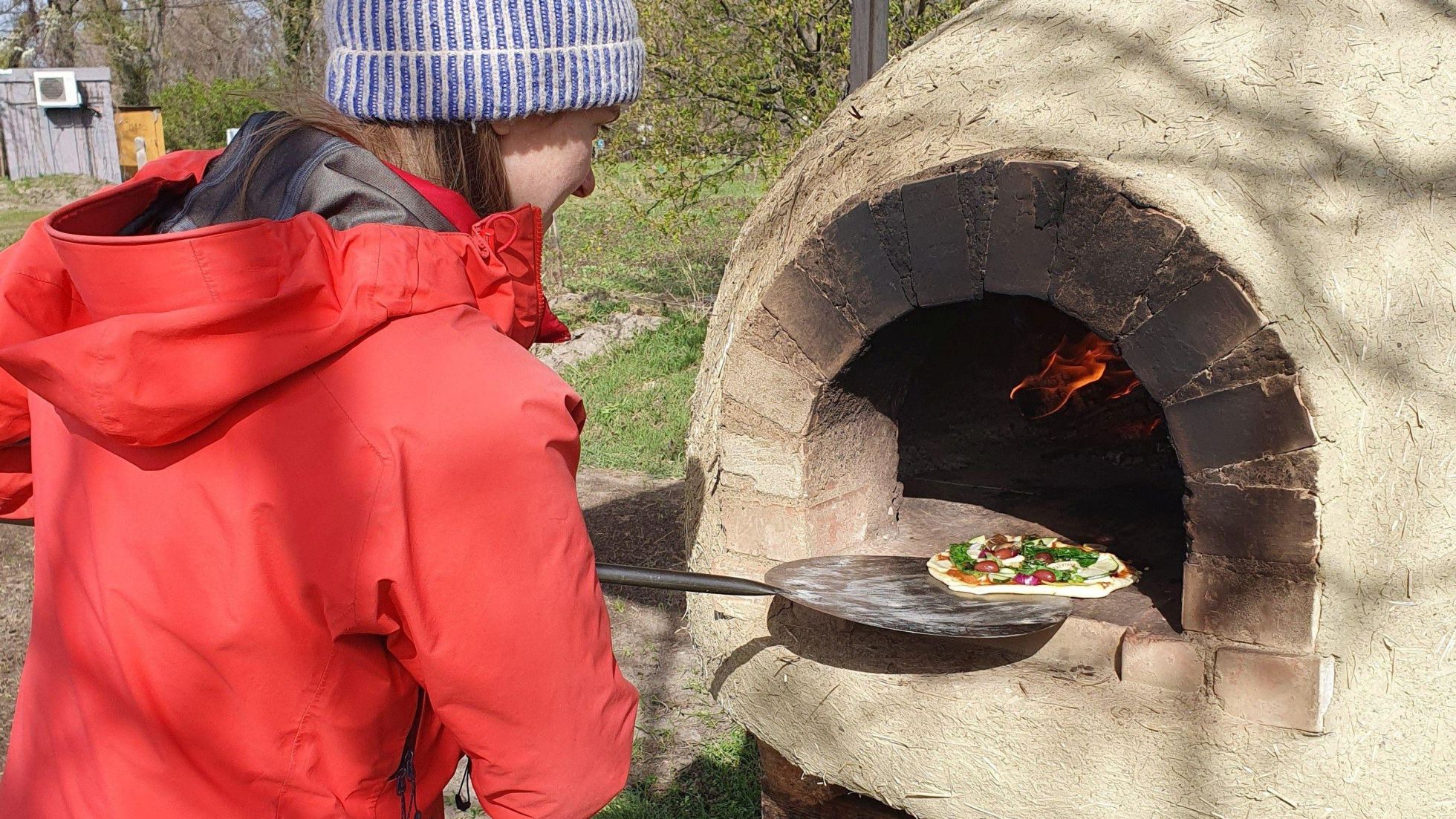 Eine Frau in einer roten Jacke und einer warmen Mütze schiebt mit einem Pizzaschieber eine Pizza in einen gemauerten Steinofen im Freien.