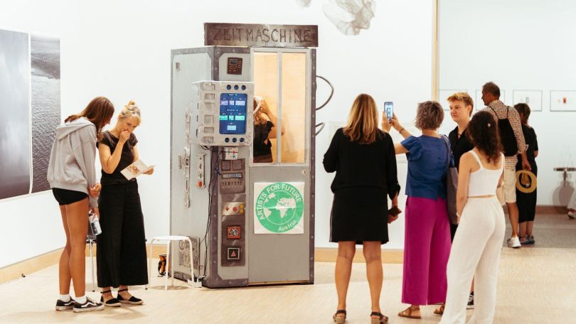 In einem Ausstellungsraum steht eine Box, die an eine alte Telefonzelle erinnert. Darüber steht "Zeitmaschine" und am Eingang klebt ein "Artists for Future"-Plakat. Rund um die Box stehen mehrere Jugendliche, eine macht davon ein Handy-Foto.