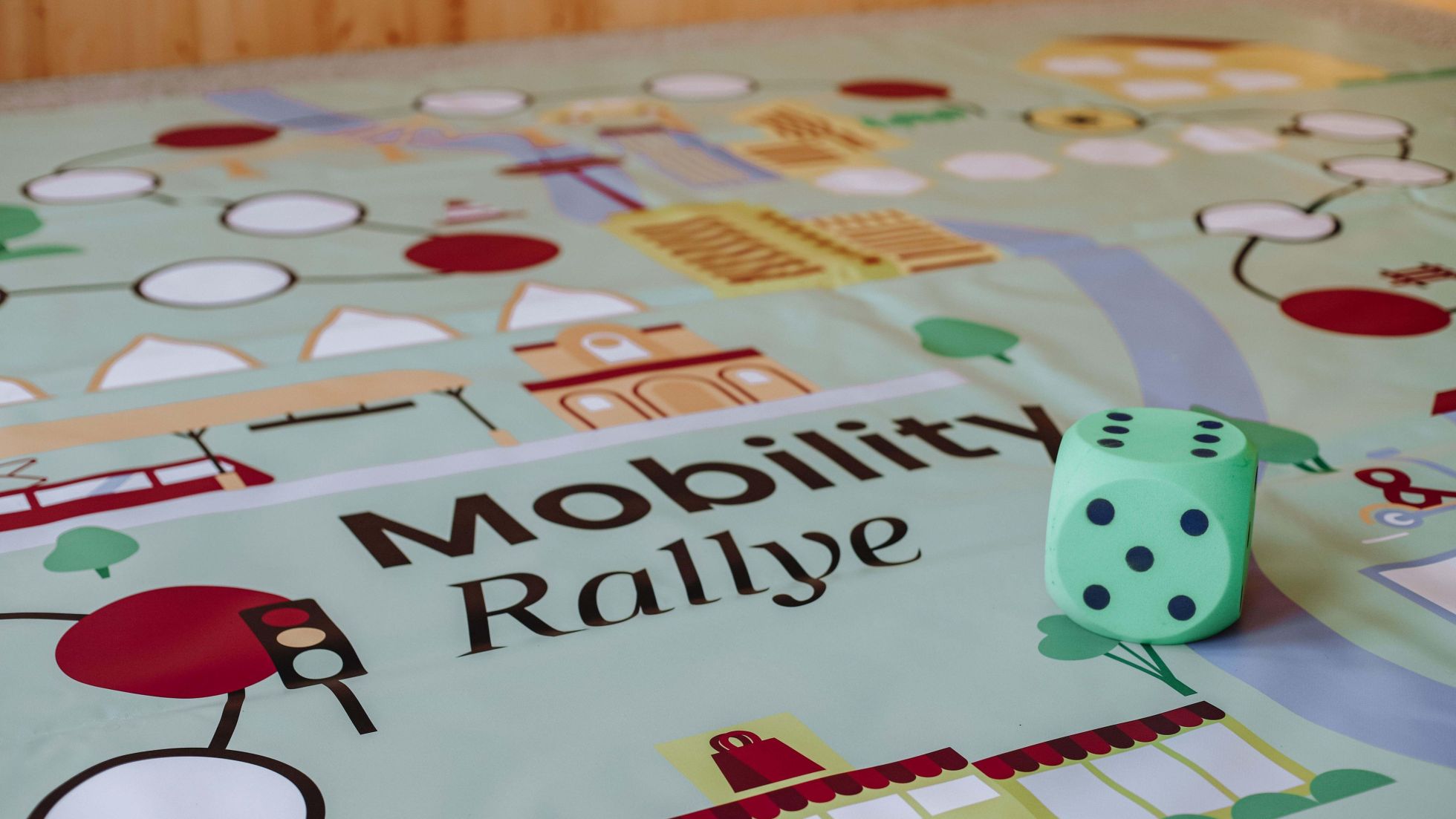 Ein hellgrüner Würfel liegt auf einem bunten Spielbrett. Am Brett steht "Mobilty Rallye".